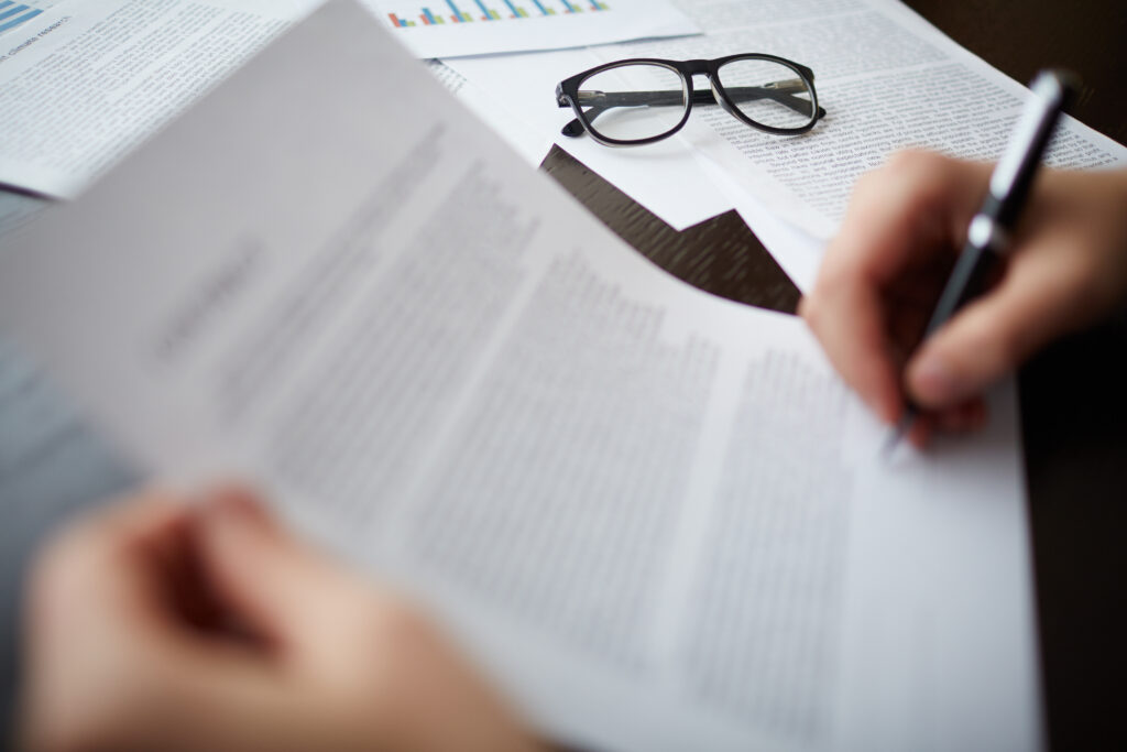 Osoba przegląda i dokonuje wpisów w wydrukowanym dokumencie, siedząc przy biurku, na którym leżą porozrzucane kartki, a obok znajdują się okulary; prawdopodobnie korzysta z materiałów takich jak „POBIERZ wzory dokumentów najmu nieruchomości”. Zdjęcie skupia się na rękach i dokumentach; twarz osoby nie jest widoczna.