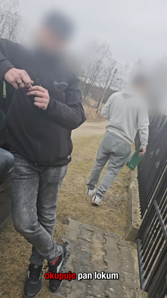 eksmisja1 Dwie osoby stoją na świeżym powietrzu w pochmurny dzień; jedna z nich opiera się o samochód ze skrzyżowanymi rękami, a jej twarz jest zamazana, podczas gdy druga odchodzi, trzymając w ręku zielony przedmiot. W tle widać czarne ogrodzenie i bezlistne drzewa. Na dole widnieje tekst w języku polskim.